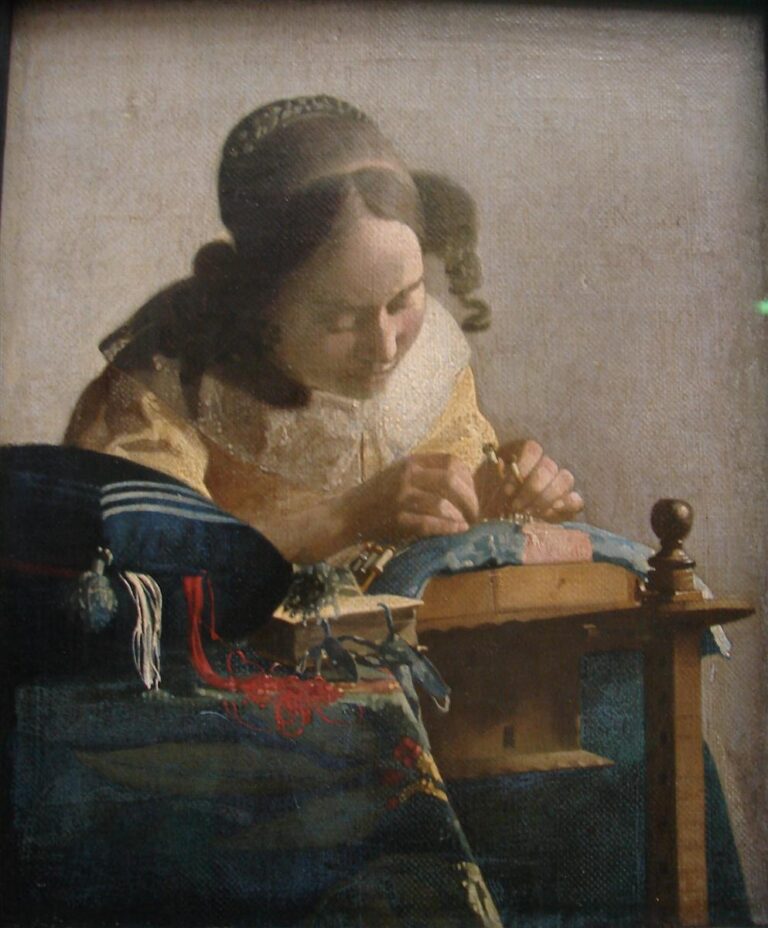 Close view of a young woman bent over a lace pillow, manipulating bobbins and thread; warm light falls on her yellow dress and white lace collar, with a blue cushion and red threads in the foreground against a plain illuminated wall.
