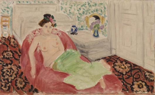 A woman reclines on a deep red couch in a patterned room; she is partially draped in a green cloth, with a dark floral carpet underfoot, a leafy wallpaper behind, and a tabletop with a vase and round mirror at the back of the scene.