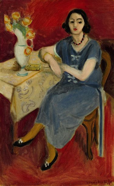 Seated woman in a blue dress with red necklace beside a table bearing a white vase of yellow flowers and a small yellow book; patterned cream tablecloth; continuous red wall and floor; mustard stockings and black shoes; calm, frontal composition with even Mediterranean light.