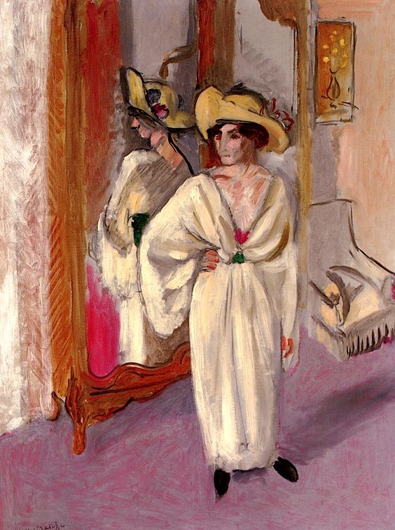 Woman in a pale white dress and wide-brimmed hat stands before a tall carved mirror in a soft mauve-and-peach room; her reflection appears in the glass as Matisse uses simplified forms, gentle brushwork, and warm–cool contrasts to explore the act of looking.