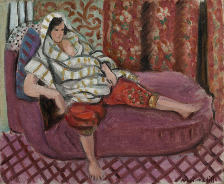 A woman reclines on a rose-colored chaise, draped in a pale patterned shawl with olive dashes and wearing russet ornamental trousers; floral curtains and a mauve lattice floor frame the scene in Matisse’s Nice-period interior.