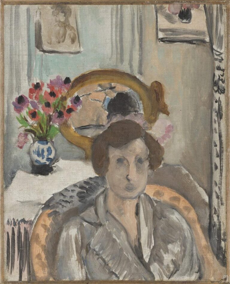 A woman in a gray robe sits in an upholstered chair before a table with a blue-and-white vase of red and pink anemones; an oval gold mirror behind her reflects head and flowers, pale curtains frame the scene, and the muted palette is enlivened by the bouquet’s bright accents.