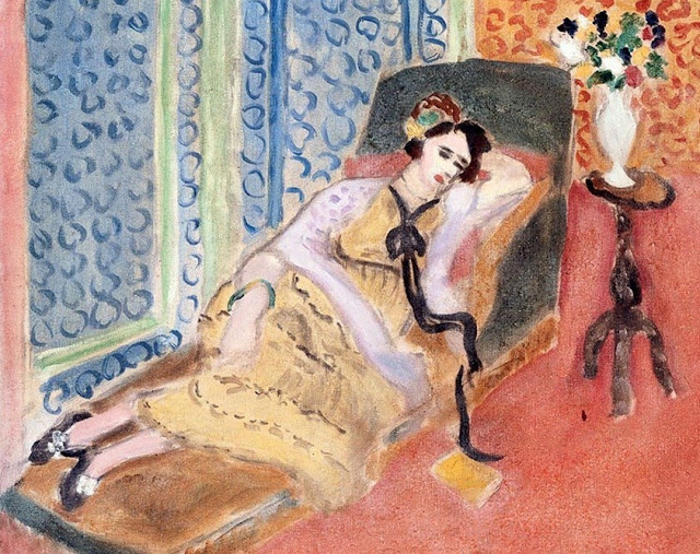 A reclining woman in a yellow dress rests on a diagonal chaise within a coral and blue patterned room; a long black ribbon falls from her collar, a white vase of flowers stands on a small table at right, and a yellow book lies on the floor, all rendered with soft, luminous color and graceful contours.