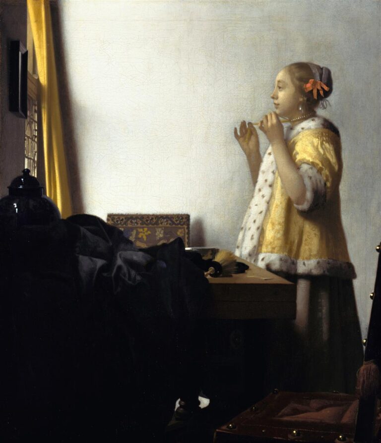 In a sunlit Delft room, a woman in a yellow fur-trimmed jacket stands in profile adjusting a pearl necklace beside a table piled with a dark cloak, a blue jar, a gilded casket, and a basin, with a yellow curtain and pale wall glowing behind her.