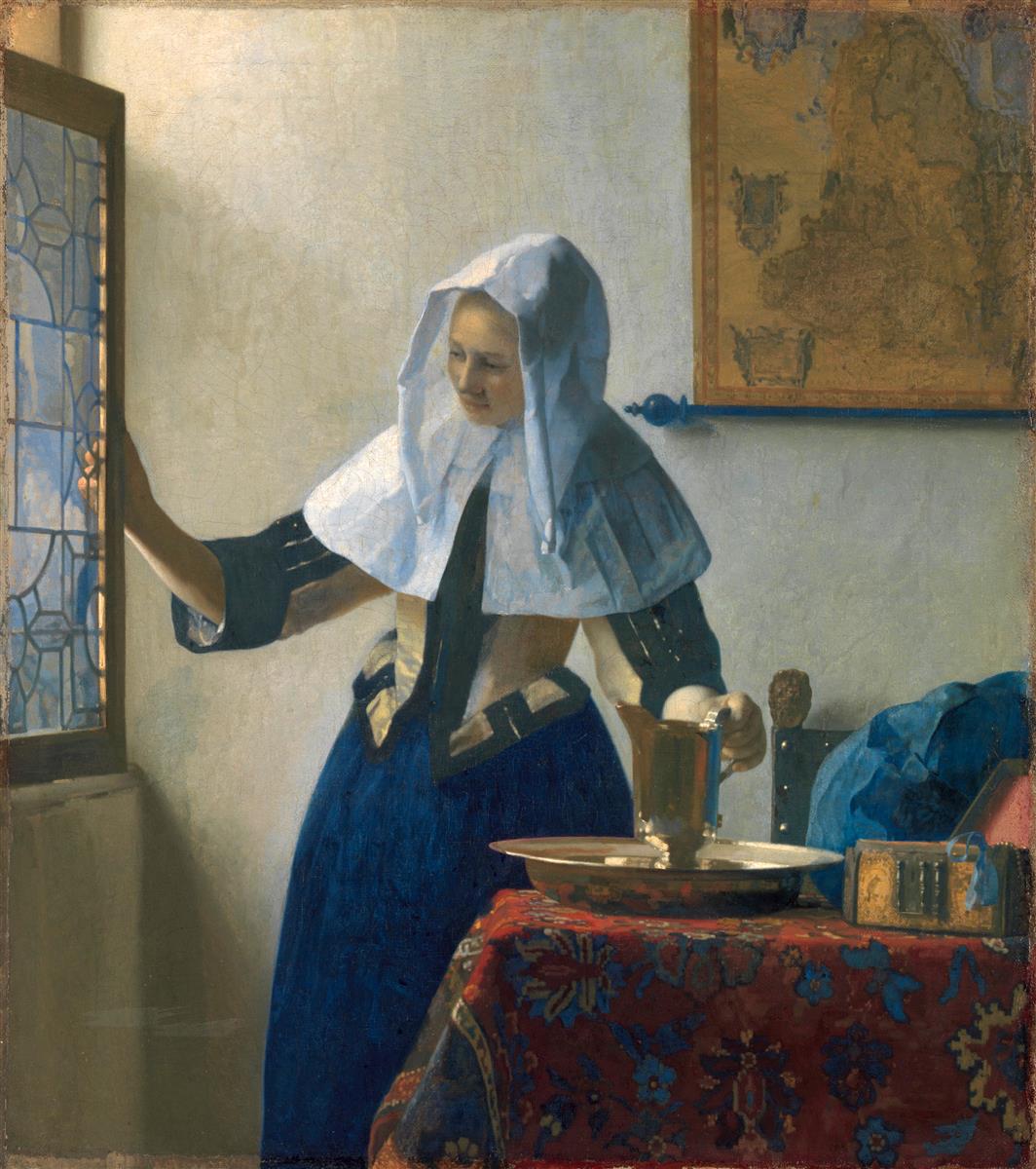 A woman in a yellow jacket and blue skirt stands by an open leaded-glass window lifting a metal water pitcher from a basin on a richly patterned table, with a blue garment, a small casket, and a wall map in a softly lit Delft room.