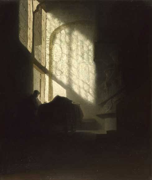 A small, dark interior with a tall arched window casting a bright lattice of light across a wall, while a solitary man sits in shadow at a table near the window.