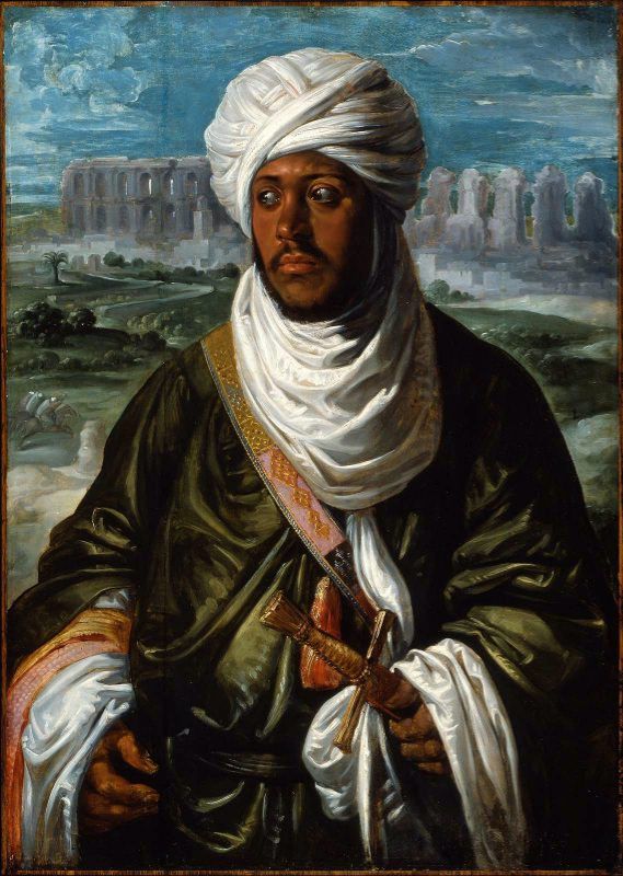 Half-length portrait of a North African ruler wearing a brilliant white turban and scarf over a dark green robe, hands resting on a dagger hilt, set before a blue sky and distant ruins in a broad landscape.
