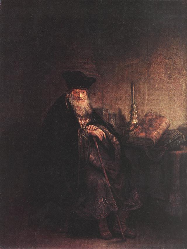 Rembrandt painting of an elderly rabbi seated in a dim room, hands folded on a staff, face and beard caught by warm light; on the table beside him lie stacked books and a tall candlestick, while the rest of the space dissolves into soft shadow.