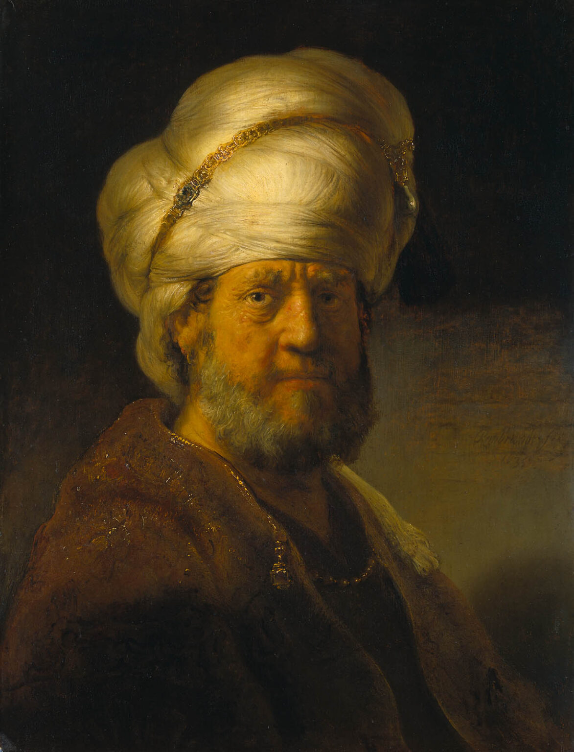 Half-length Rembrandt portrait of an older man wearing a large white turban and brown robe with a gold chain, his bearded face lit against a dark background, emphasizing rich textures and a steady, thoughtful gaze.
