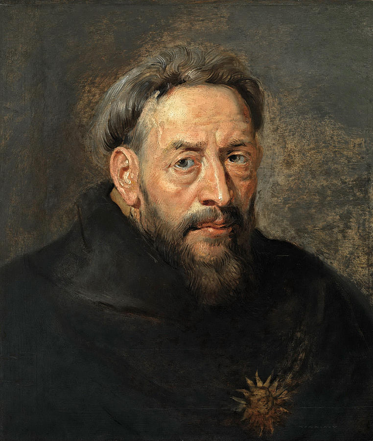 Close-up portrait of a middle-aged Capuchin monk in a dark brown habit, with graying hair, beard, and thoughtful eyes, set against a muted brown-gray background with a small golden sunburst emblem on his chest.