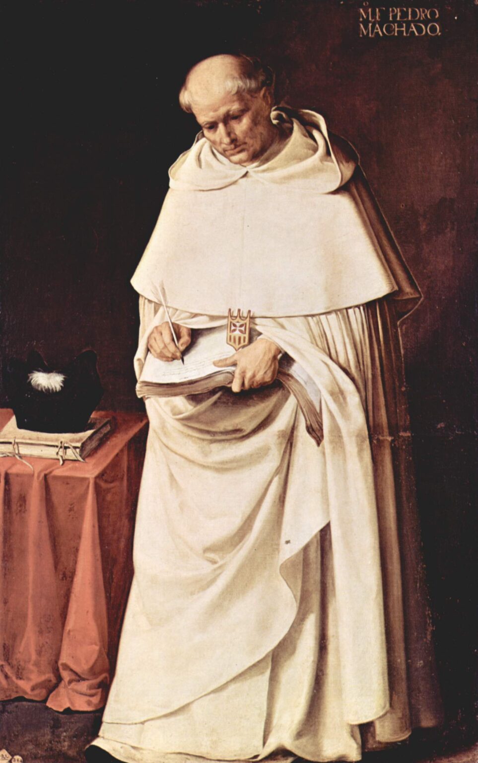 A Mercedarian monk in a voluminous white habit stands against a dark background, head bowed as he writes with a quill in a large book held in his hands, while a small red draped table beside him supports two closed books and a black clerical hat.